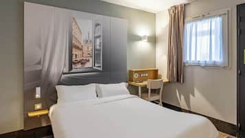 A hotel room with a large bed, a desk with a chair, a wall-mounted TV, and a window with curtains.