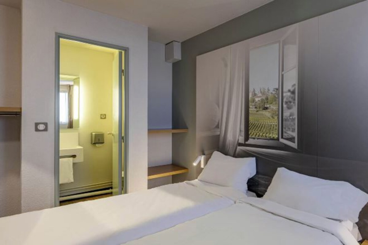 A modern hotel room with a bed, a window with a view, a door leading to a bathroom, and a small shelf.
