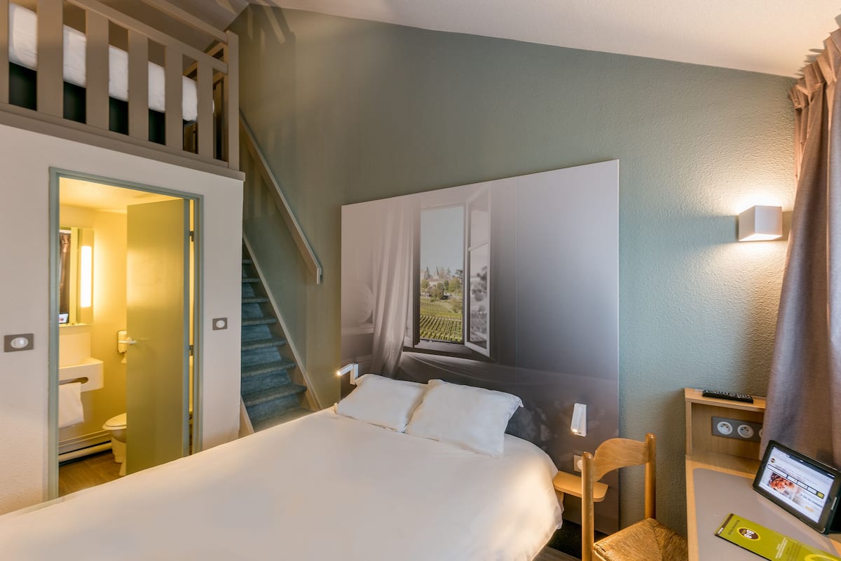 A hotel room with a bed, a desk with a computer, a window with curtains, a staircase, and a bathroom.