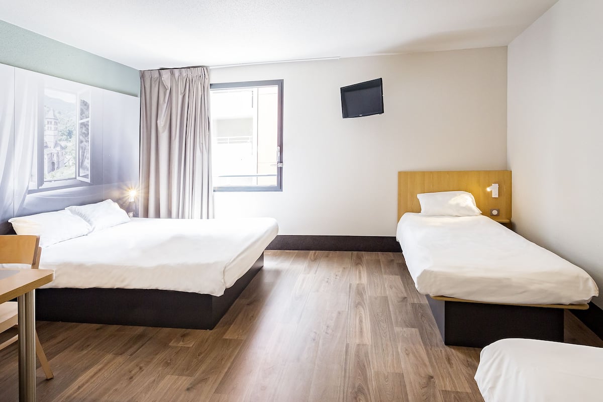 A hotel room with two beds, a desk, a window with curtains, and a flat-screen TV on the wall.