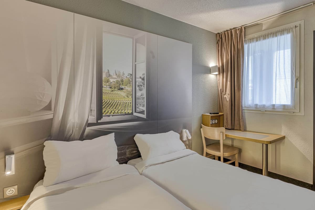 A hotel room with two beds, a desk, a chair, and a window with curtains.