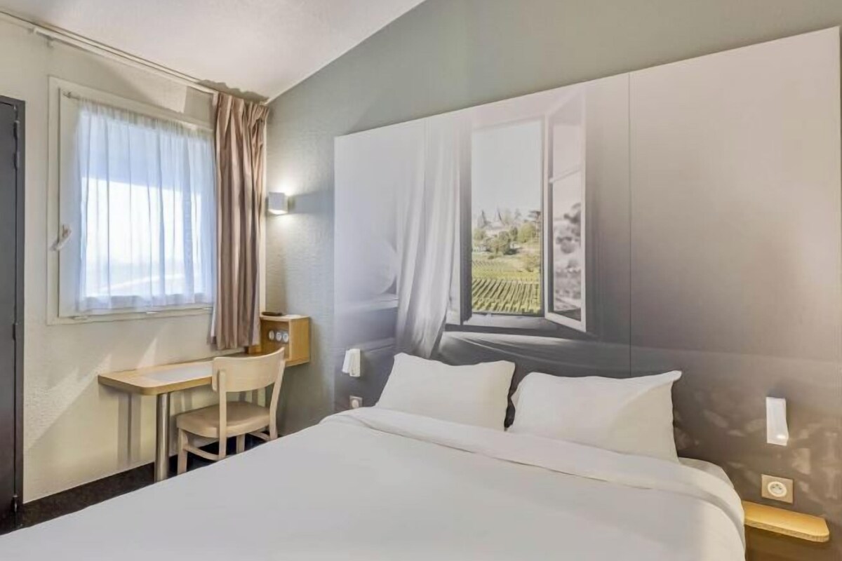 Ett hotellrum med en säng, ett skrivbord, en stol, ett fönster med gardiner och en väggmonterad lampa.