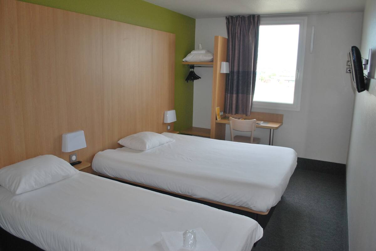A hotel room with two single beds, a desk, a chair, a window with curtains, and a wall-mounted phone.
