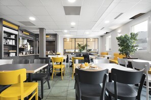 A modern cafeteria with yellow and black chairs, tables, and a kitchen area with shelves and appliances. - B&B HOTEL Lyon Vénissieux (Venissieux)