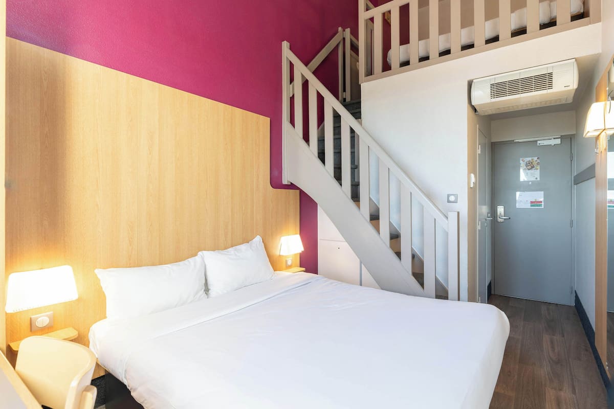A hotel room with a bed, a nightstand with a lamp, a wooden headboard, a staircase, and a door with a light switch.
