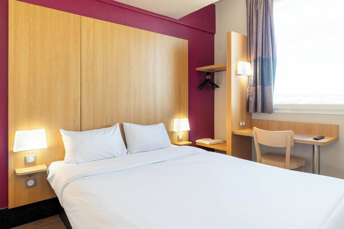 A hotel room with a large bed, bedside lamps, a desk with a chair, a mirror, and a window with curtains.