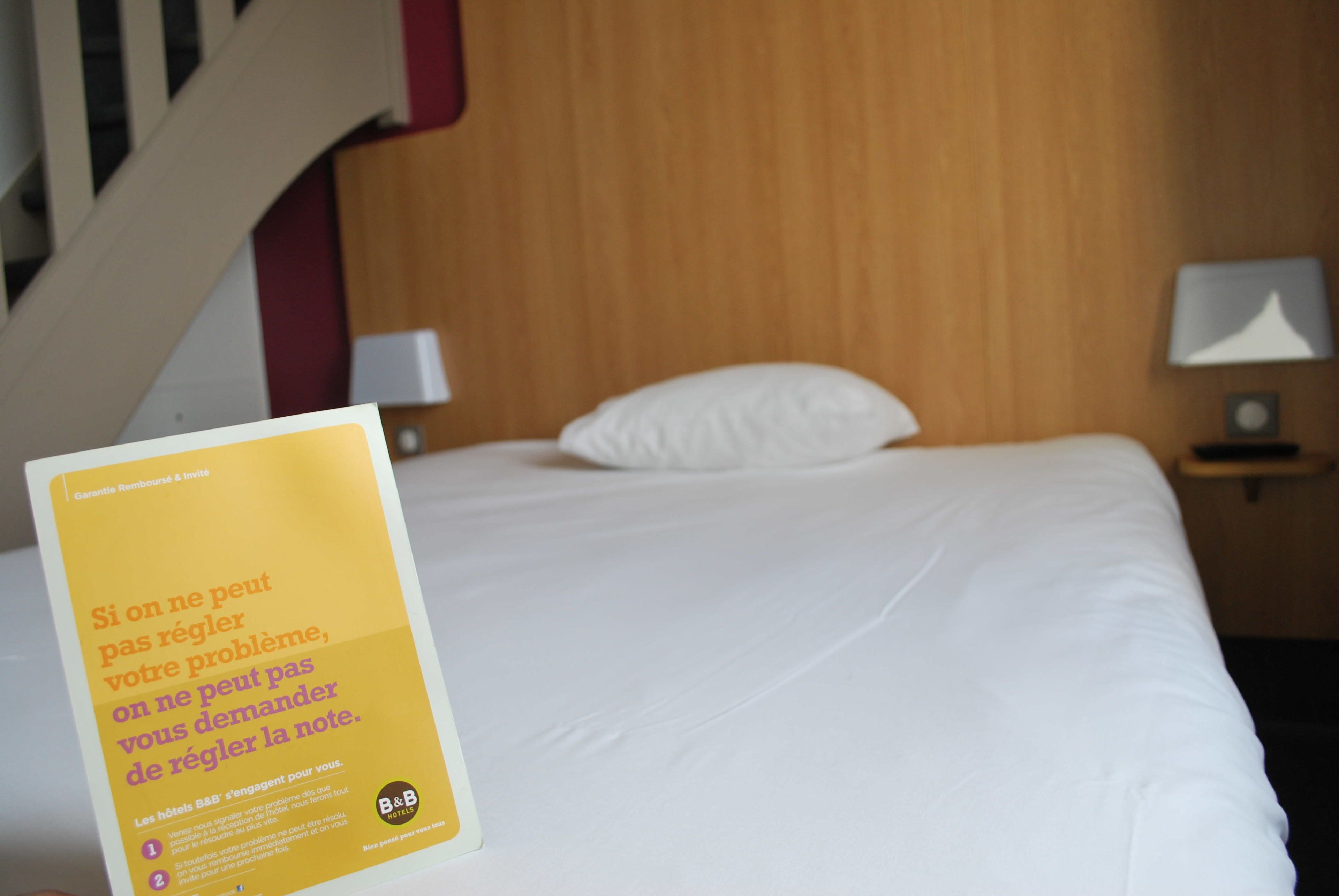 A hotel room with a bed, a wooden headboard, a nightstand with a lamp, and a sign on the bed.