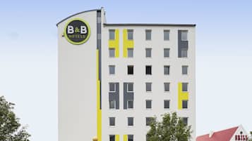 A B&B hotel with a modern design, featuring a prominent logo on the facade and a red-roofed building adjacent to it.