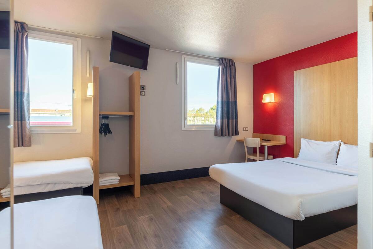 A hotel room with a large bed, two smaller beds, a TV, a desk, and a red accent wall.