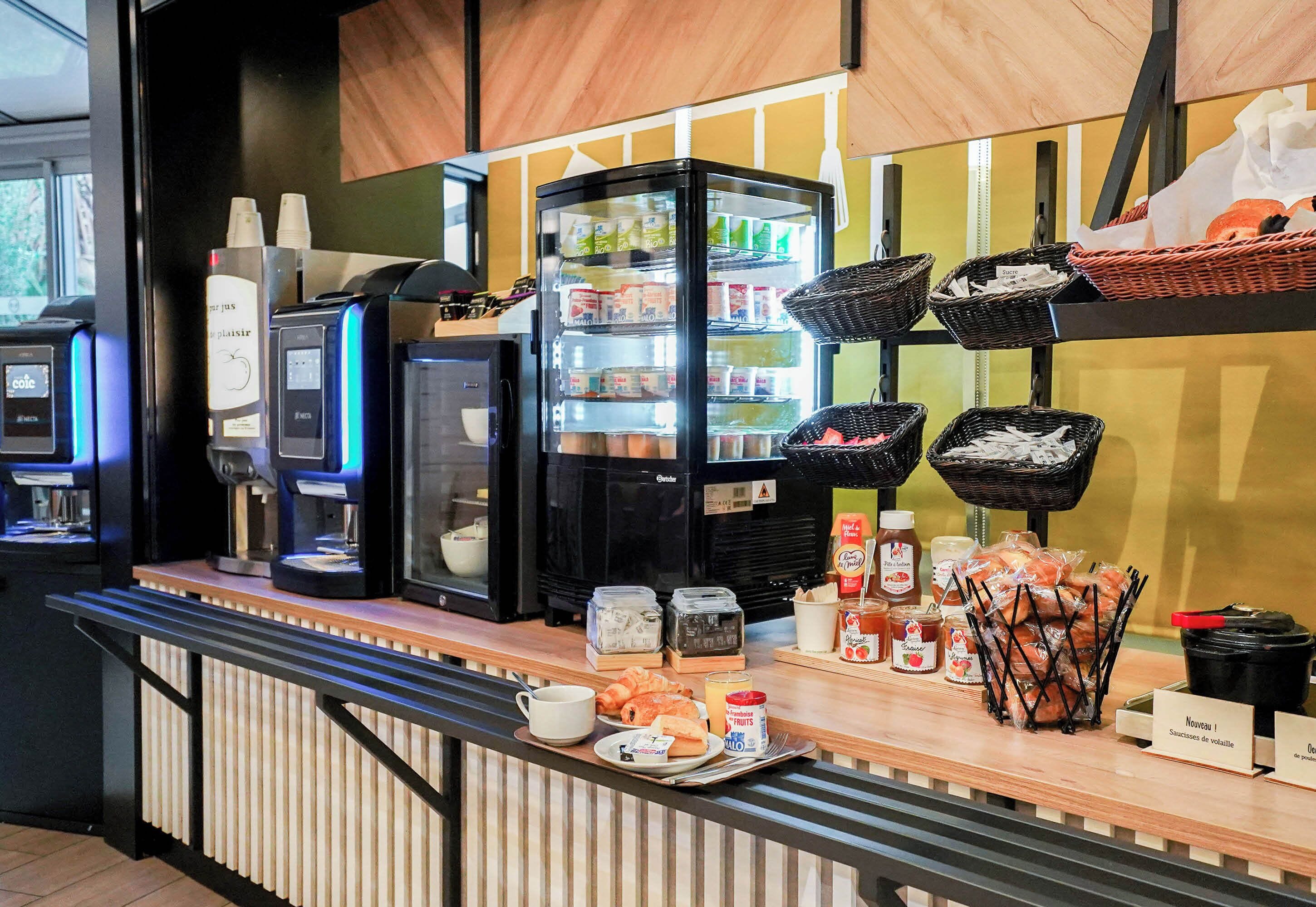A breakfast bar with coffee machines, a refrigerator stocked with beverages, baskets of bread, and various breakfast items.