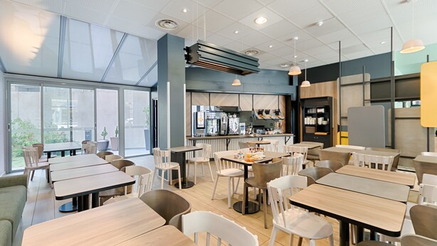 A modern cafe with wooden tables and white chairs, large windows, and a kitchen area. - B&B HOTEL Marseille Centre La Joliette (Marseille)