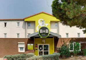 L’entrée d’un hôtel avec une façade jaune, une enseigne indiquant « B&B Hotels » et une petite fenêtre avec un store blanc.
