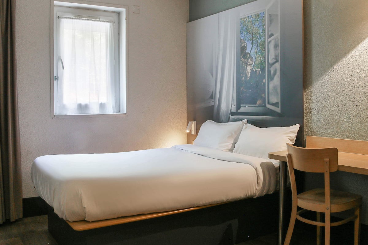 Ein ordentlich bezogenes Bett mit weißen Leinen, ein Nachttisch aus Holz, ein Holzstuhl und ein Fenster mit durchsichtigen Vorhängen.