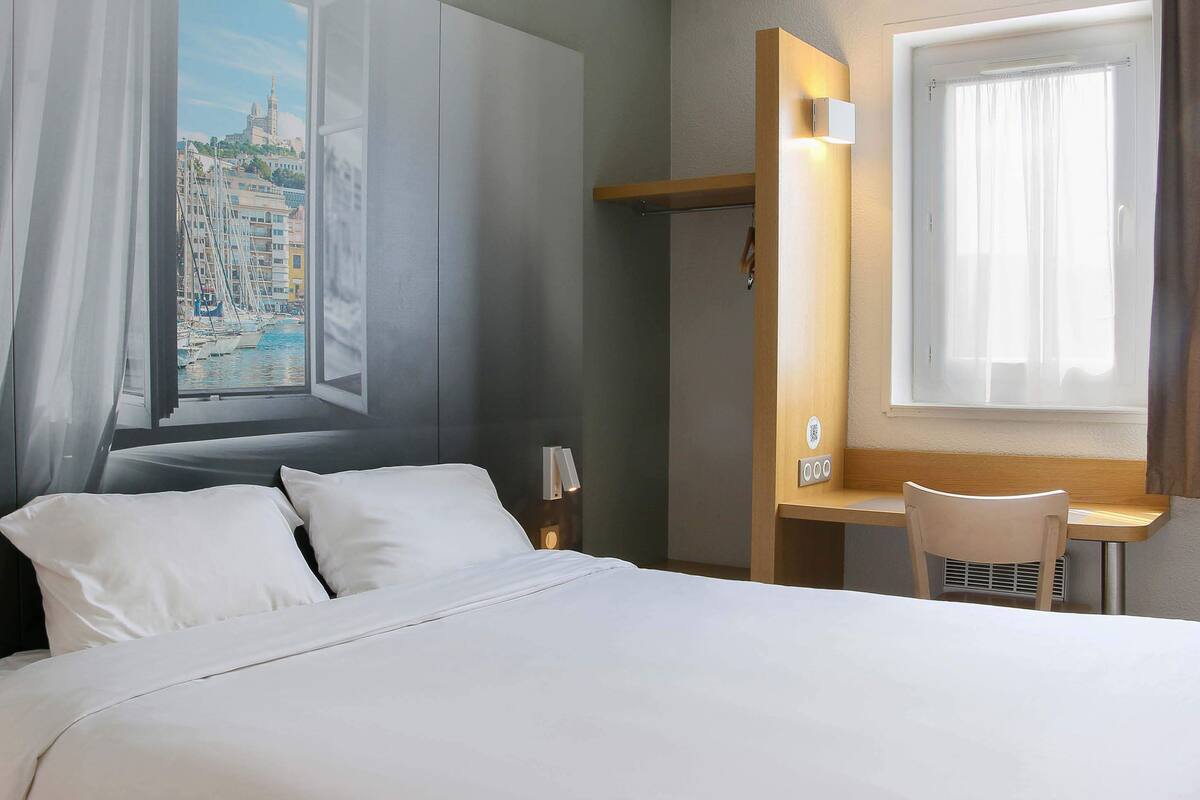 Ein Hotelzimmer mit Bett, Schreibtisch, Stuhl, einem Fenster mit Vorhängen und Blick auf eine Stadtlandschaft.
