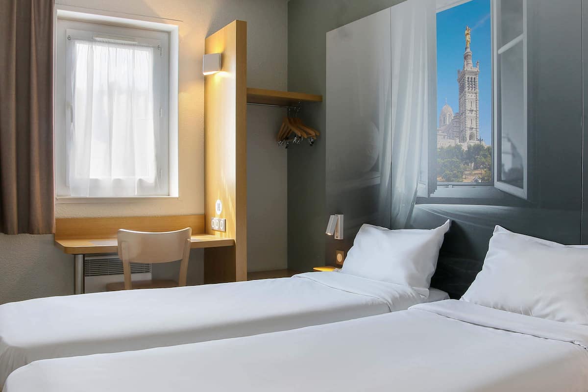 Ein Hotelzimmer mit zwei Betten, einem Schreibtisch, einem Stuhl und einem großen Fenster mit Blick auf ein Gebäude.
