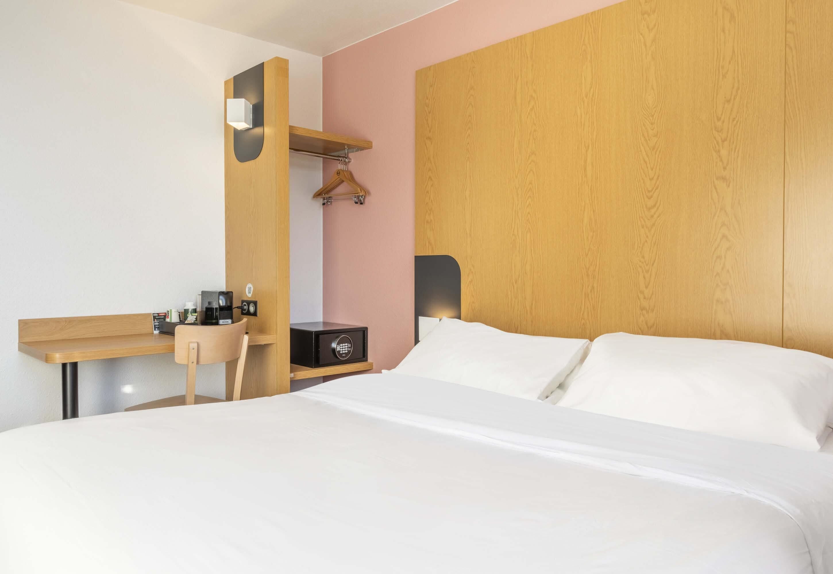 A hotel room with a bed, bedside table, a chair, a hanging coat rack, and a wooden headboard.