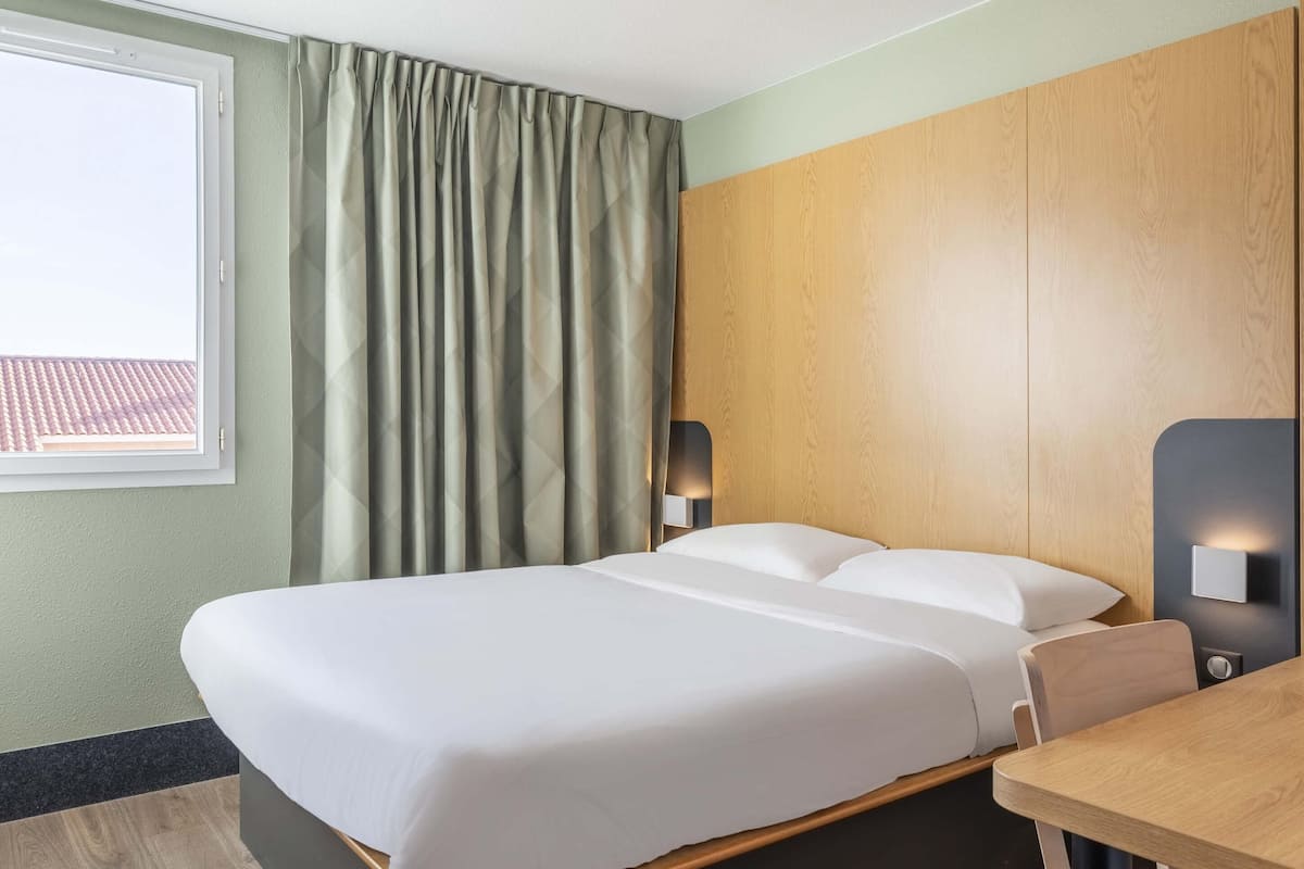 A hotel room with a bed, a wooden headboard, a table, and a window with curtains.