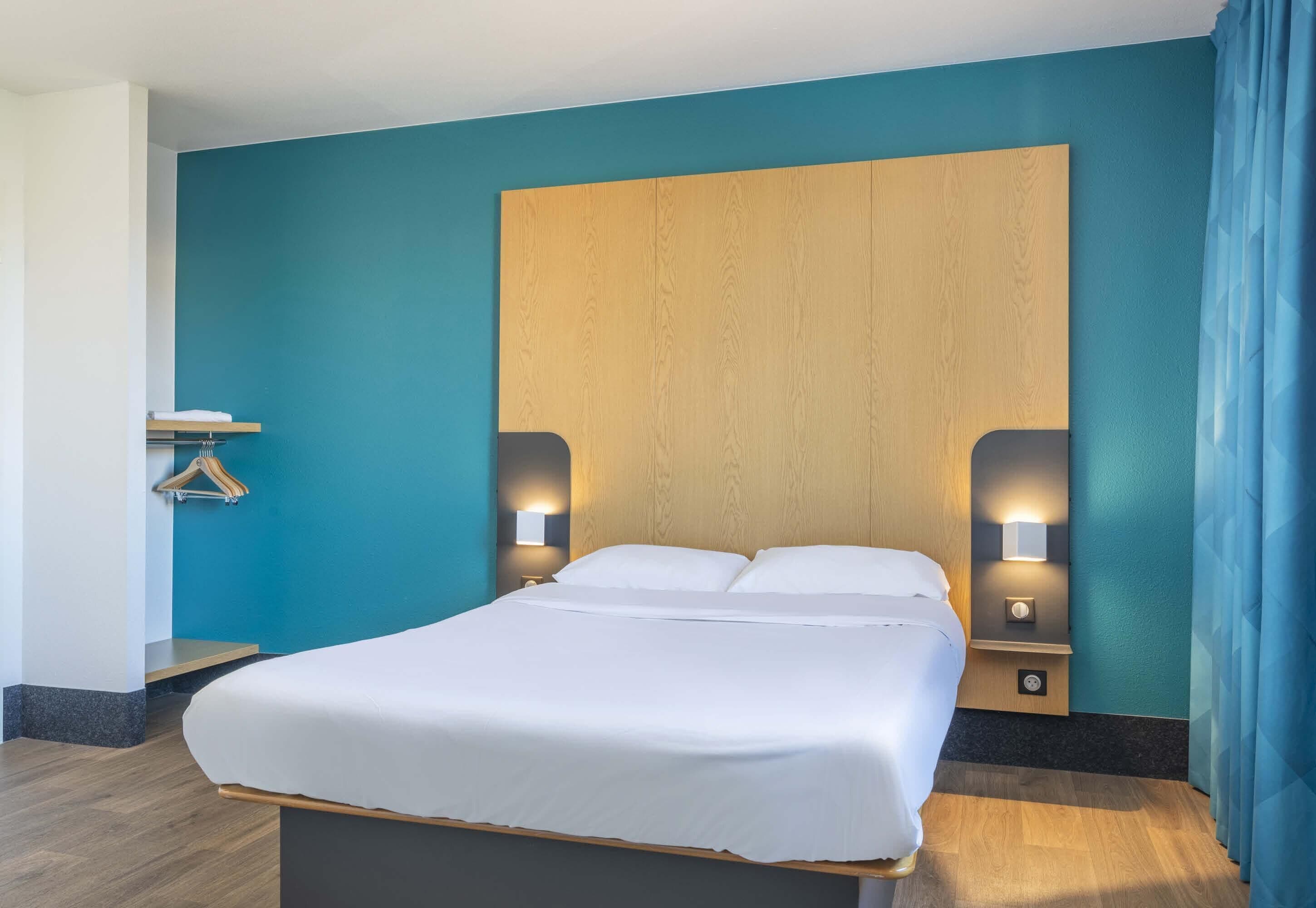 A modern hotel room with a large bed, wooden headboard, bedside lamps, and a blue accent wall.