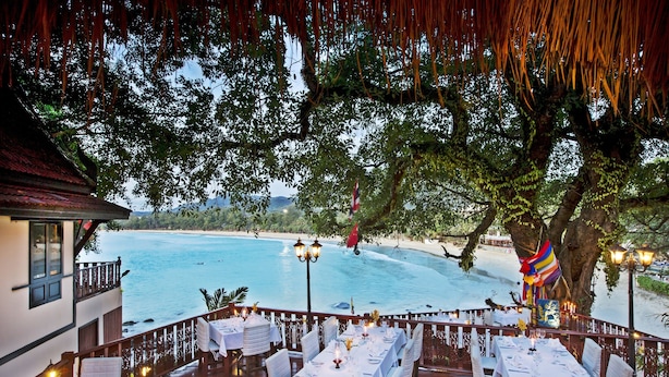 Un área de comedor al aire libre con mesas dispuestas para una comida, con vistas a una playa y al mar.