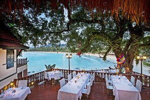 An outdoor dining area with tables set for a meal, overlooking a beach and the sea.