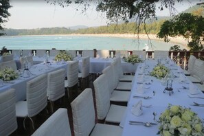 Una larga mesa de comedor dispuesta para una boda, con sillas blancas y centros de mesa florales, con vistas a una playa.