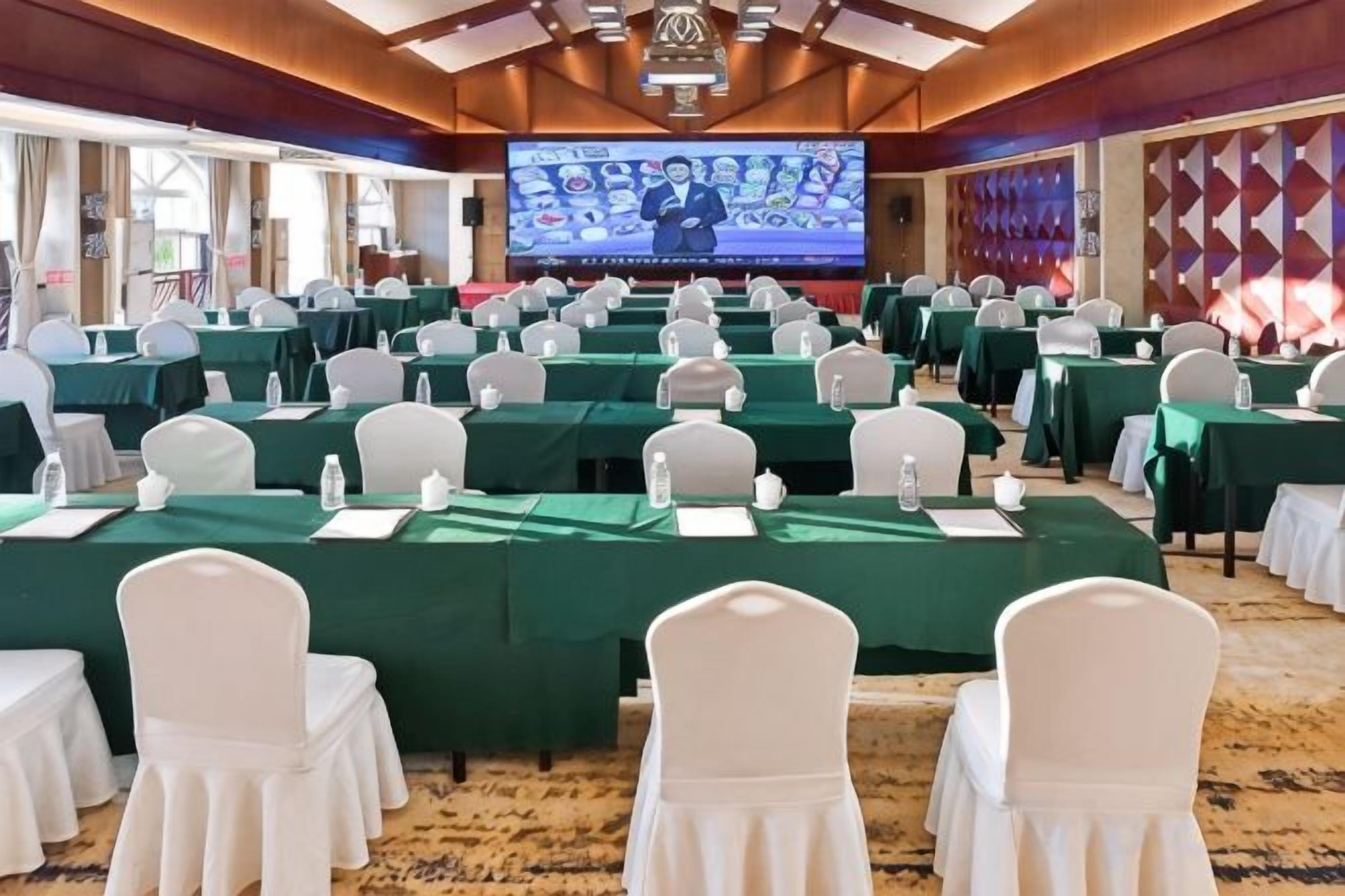 A conference room with rows of tables and chairs, a large screen at the front, and a carpeted floor.