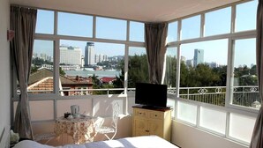 Ein Schlafzimmer mit einem großen Bett, einem Fernseher auf einer Kommode, einem kleinen Tisch mit einem Stuhl und einem Balkon mit Blick auf die Stadt.