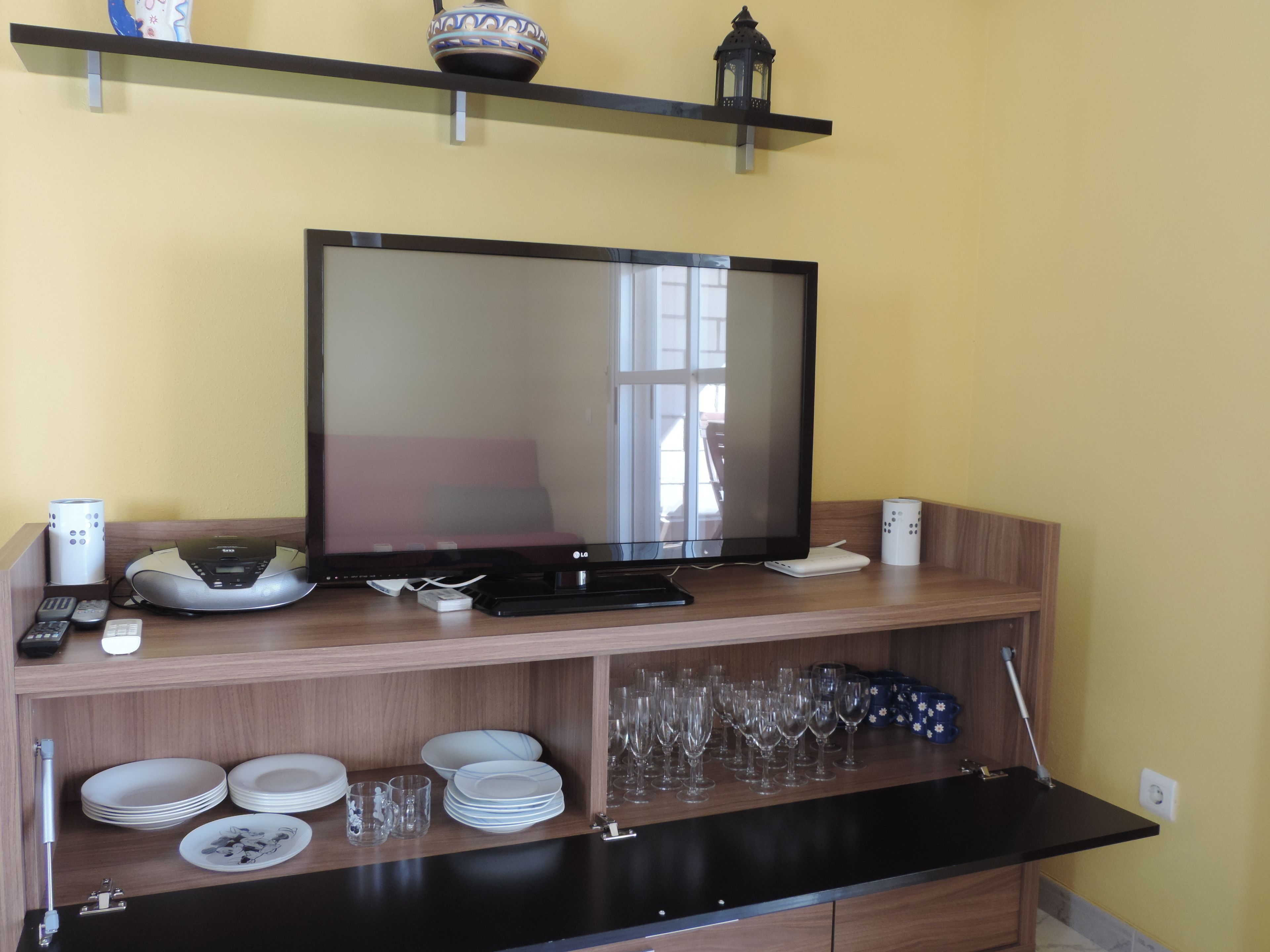 Un televisor de pantalla plana sobre un soporte de madera, con un control remoto, una balanza y un dispositivo blanco a la izquierda. Abajo, se apilan platos, vasos y un recipiente azul.