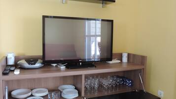 Un televisor de pantalla plana sobre un soporte de madera, con un control remoto, una balanza y un dispositivo blanco a la izquierda. Abajo, se apilan platos, vasos y un recipiente azul.