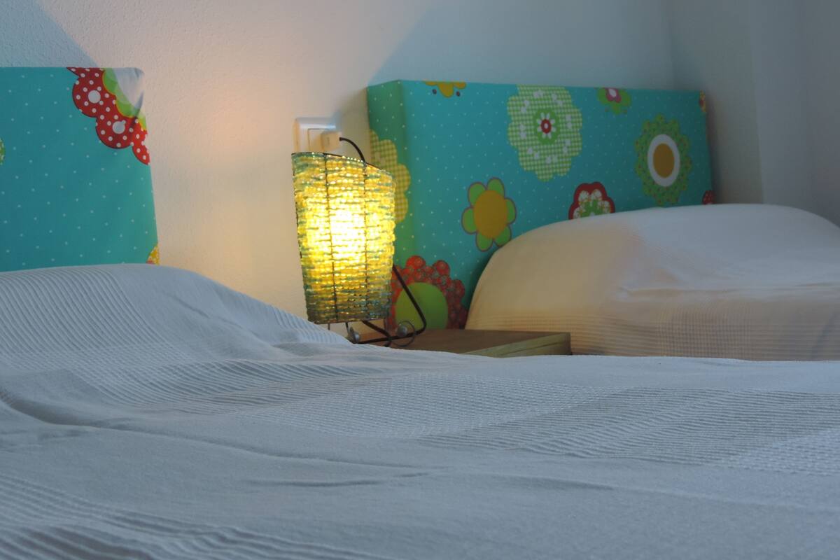 Ein Bett mit weißer Bettwäsche, ein Nachttisch mit Lampe und ein farbenfroher Kopfteil.