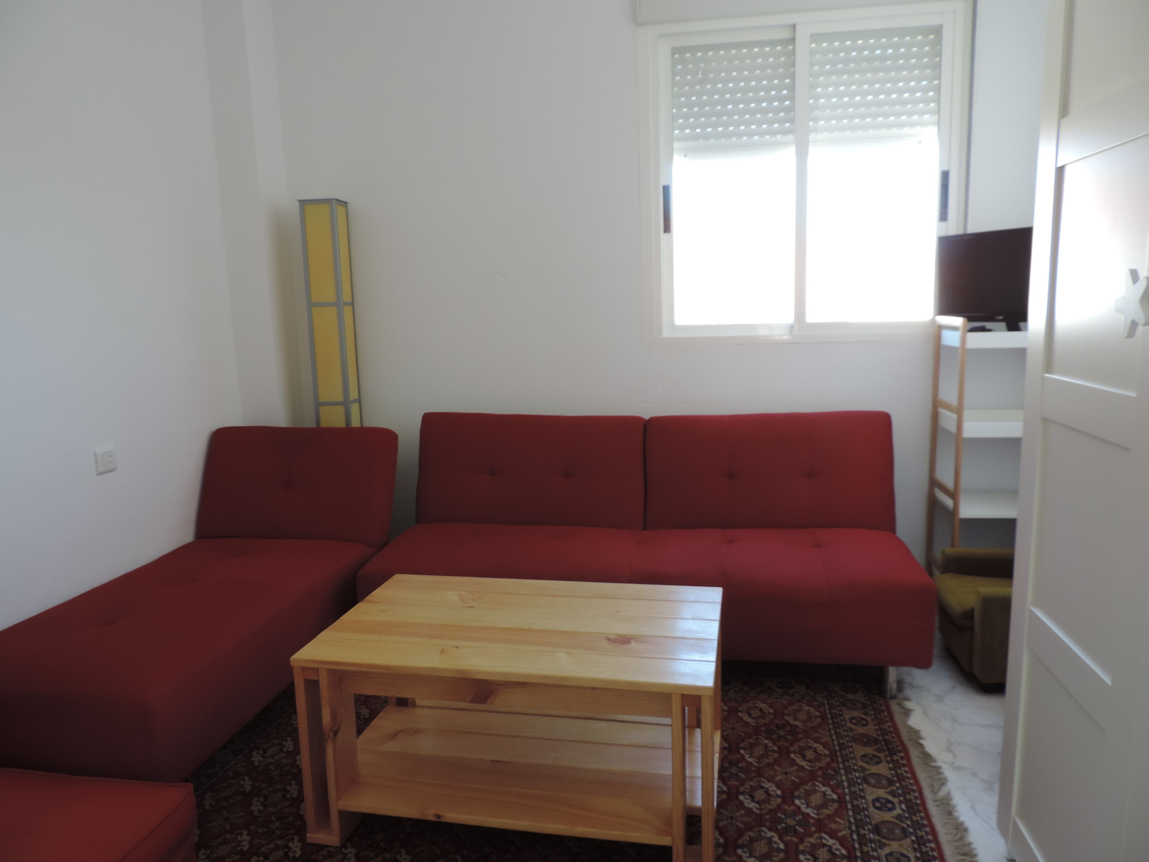 Un petit salon avec un canapé rouge, une table basse en bois et une fenêtre équipée de stores.