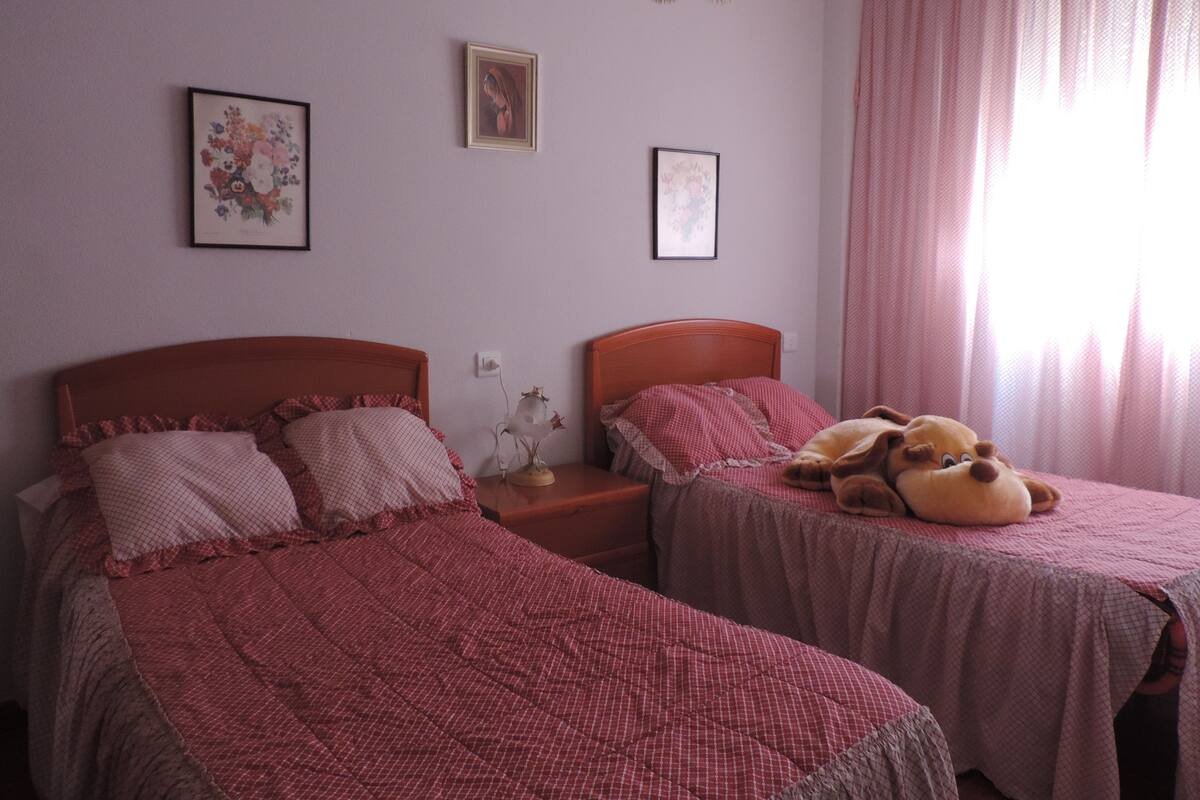 Ein Schlafzimmer mit zwei Betten, einem Nachttisch, einer Lampe und einem Fenster mit Vorhängen.