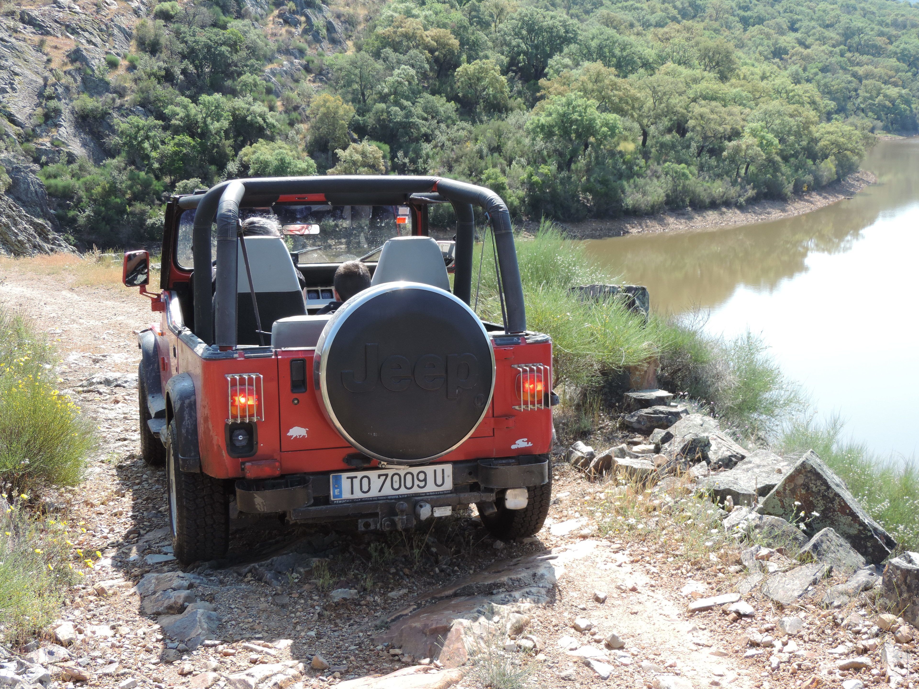 A red Jeep with a spare tire mounted on the back, parked on a rocky path beside a river.