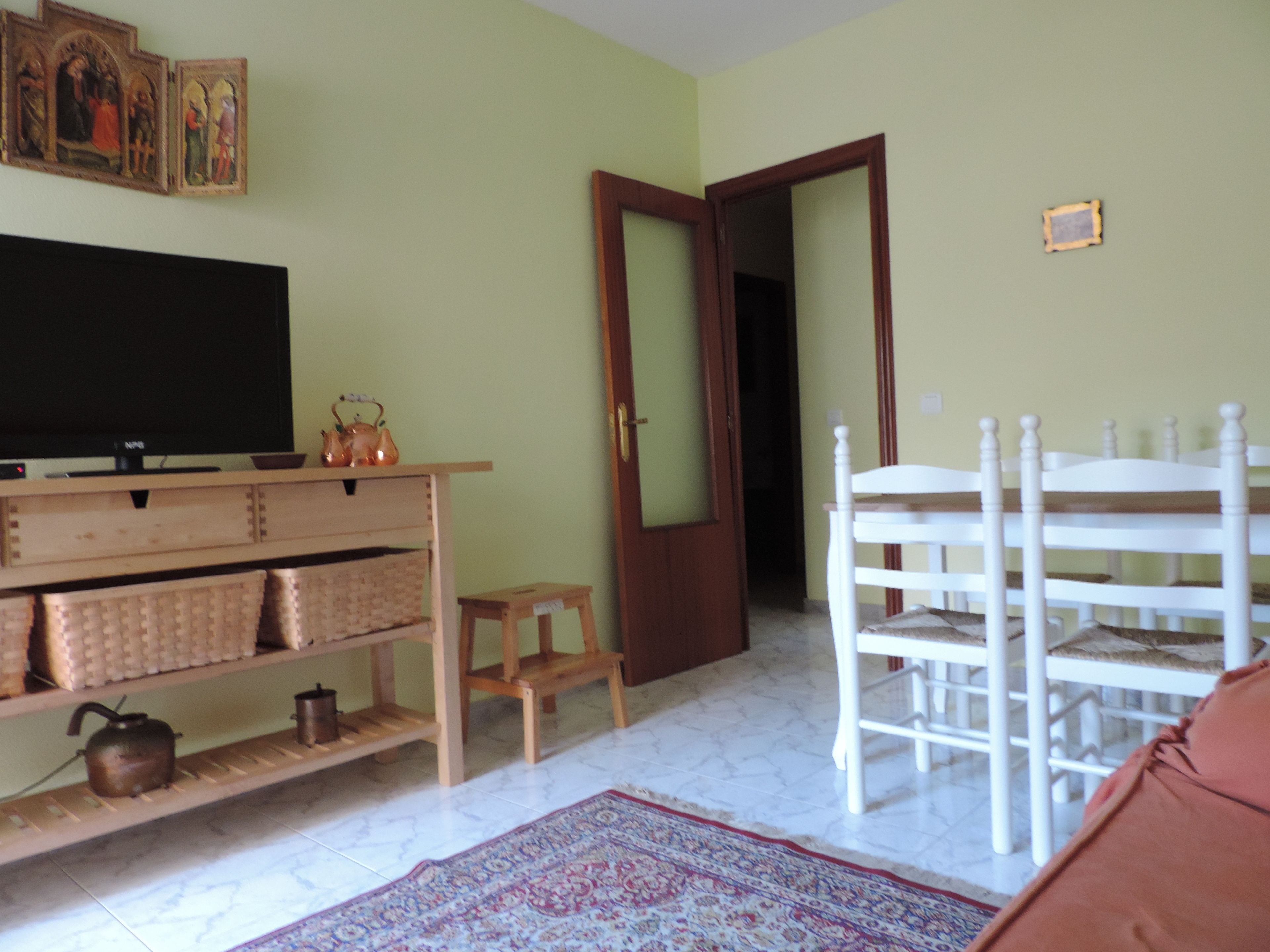 Una sala de estar con televisor, muebles de madera, un tapete estampado y una puerta que da a otra habitación.