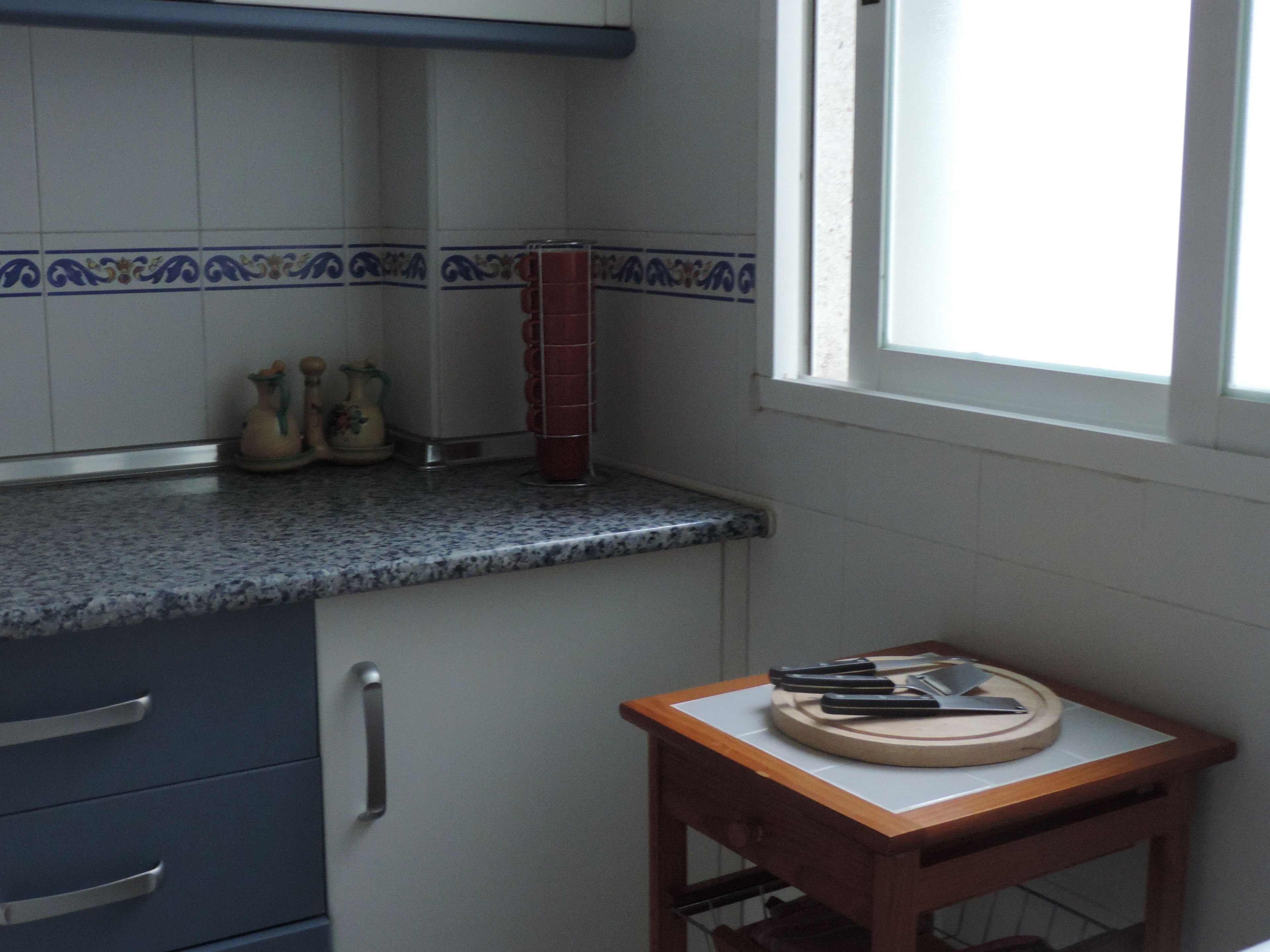 Een keuken met een granieten aanrecht, een kleine houten tafel met een snijplank en een raam met jaloezieën.