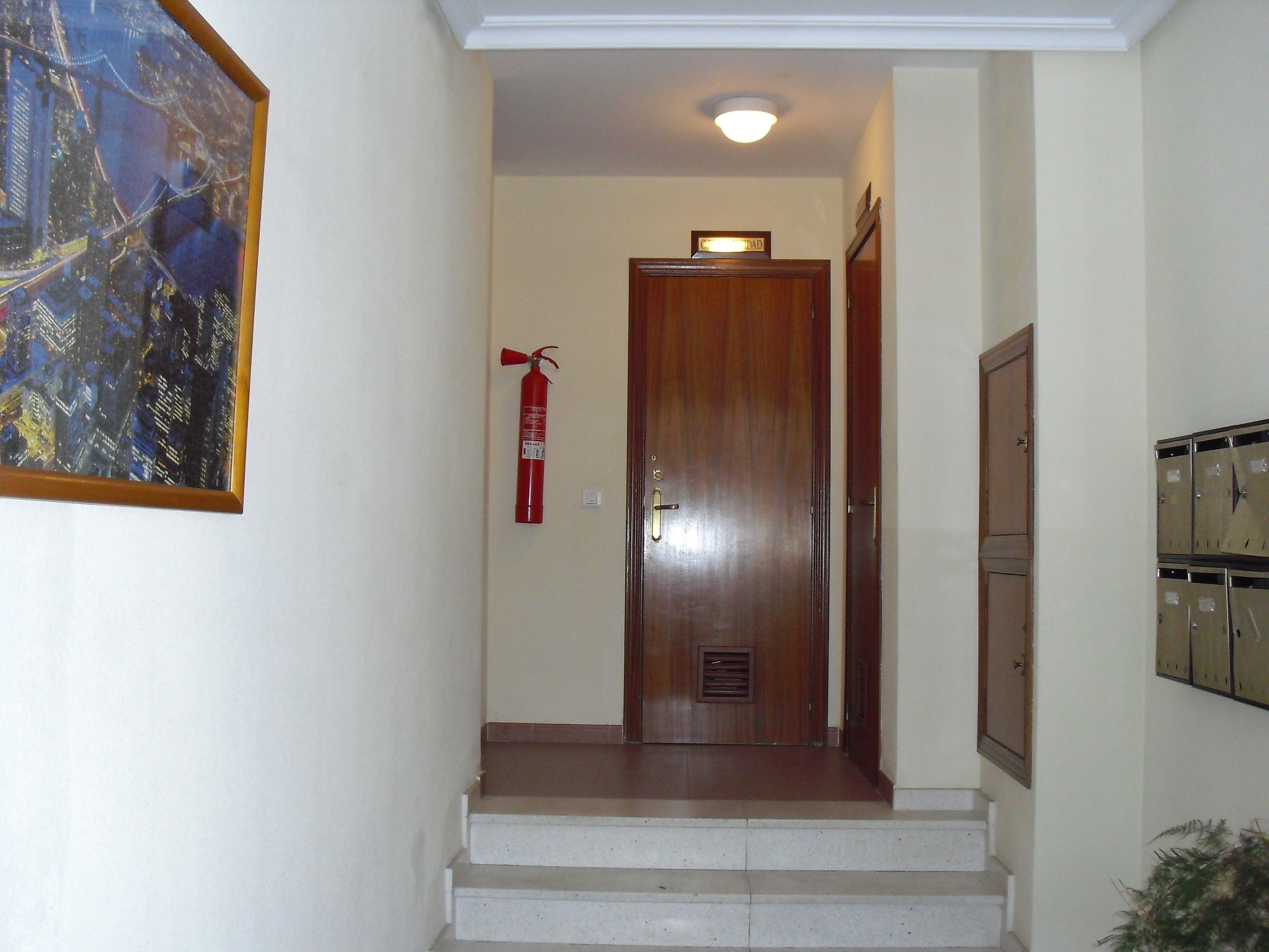 Un pasillo con un extintor de incendios montado en la pared, una puerta de madera y una serie de taquillas a la derecha.
