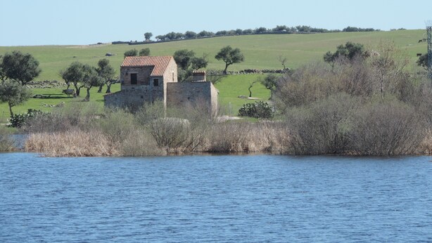 A stone building with a tiled roof, surrounded by a grassy field and trees, near a body of water. - Apartamentos Barbancho (Casar de Caceres)