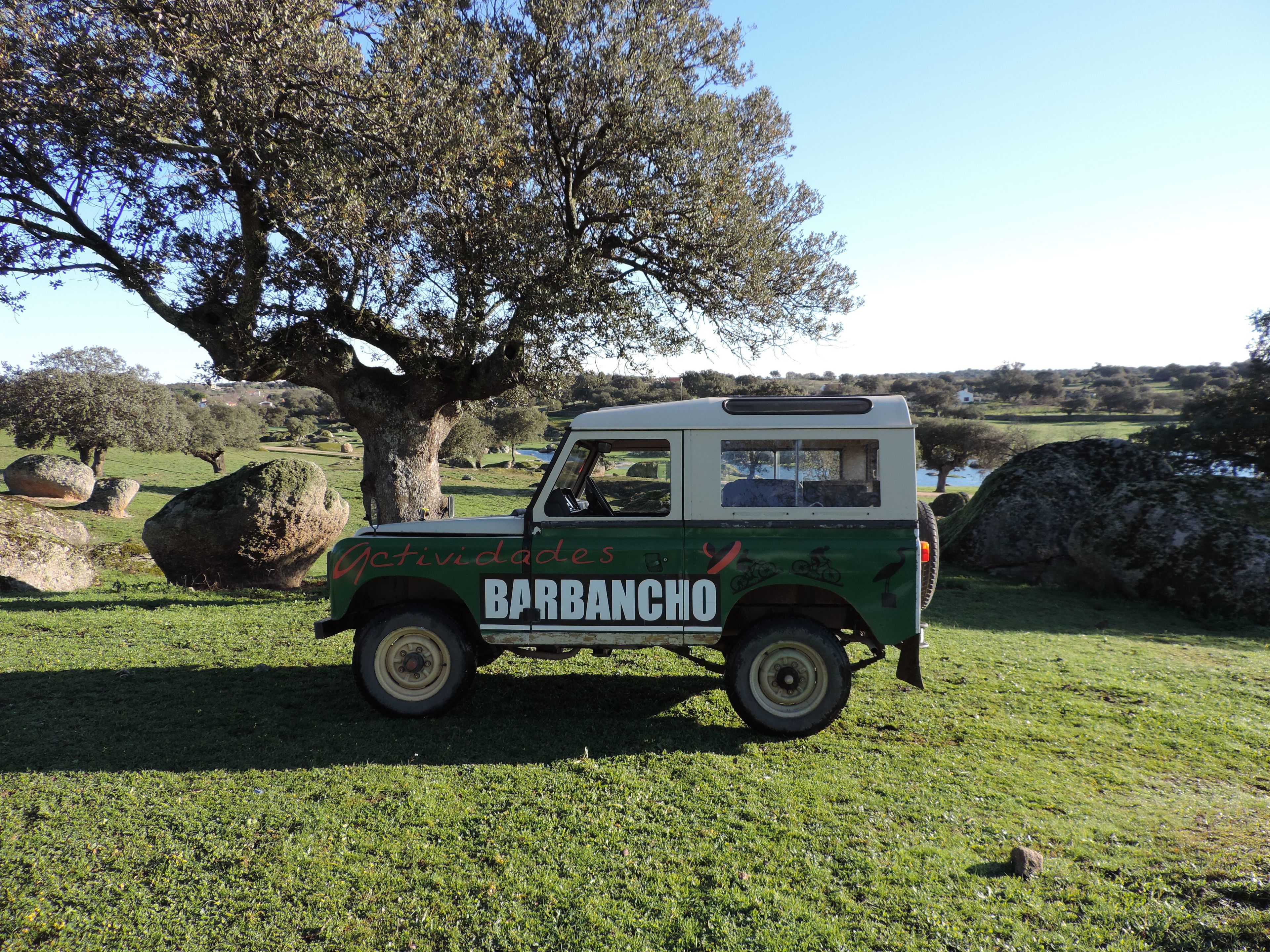 Ein grün-weißes Fahrzeug mit der Aufschrift "Jctividades BARBANCHO", geparkt in einer grünen Umgebung mit großen Steinen und einem Baum im Hintergrund.