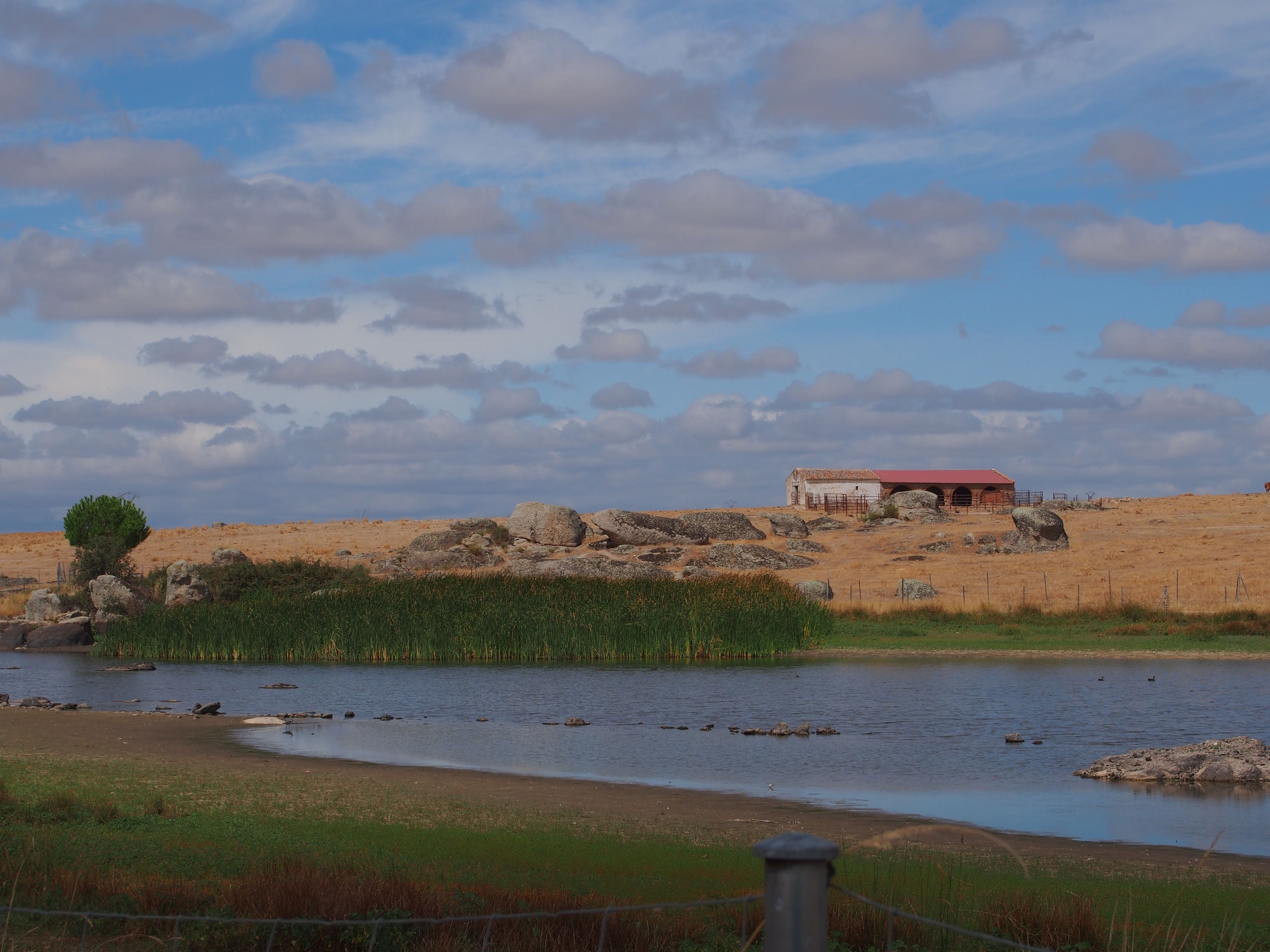 Un paisaje rural con un río, un pequeño edificio de techo rojo y una valla.