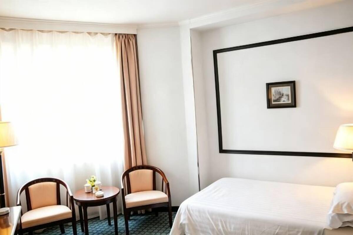 Hotellihuone, jossa on sänky, kaksi tuolia, pieni pöytä, lamppu ja seinällä oleva kehystetty kuva.