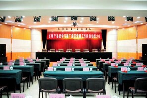 Un salón de conferencias con filas de mesas y sillas, un escenario con un telón de fondo rojo, y un cartel.