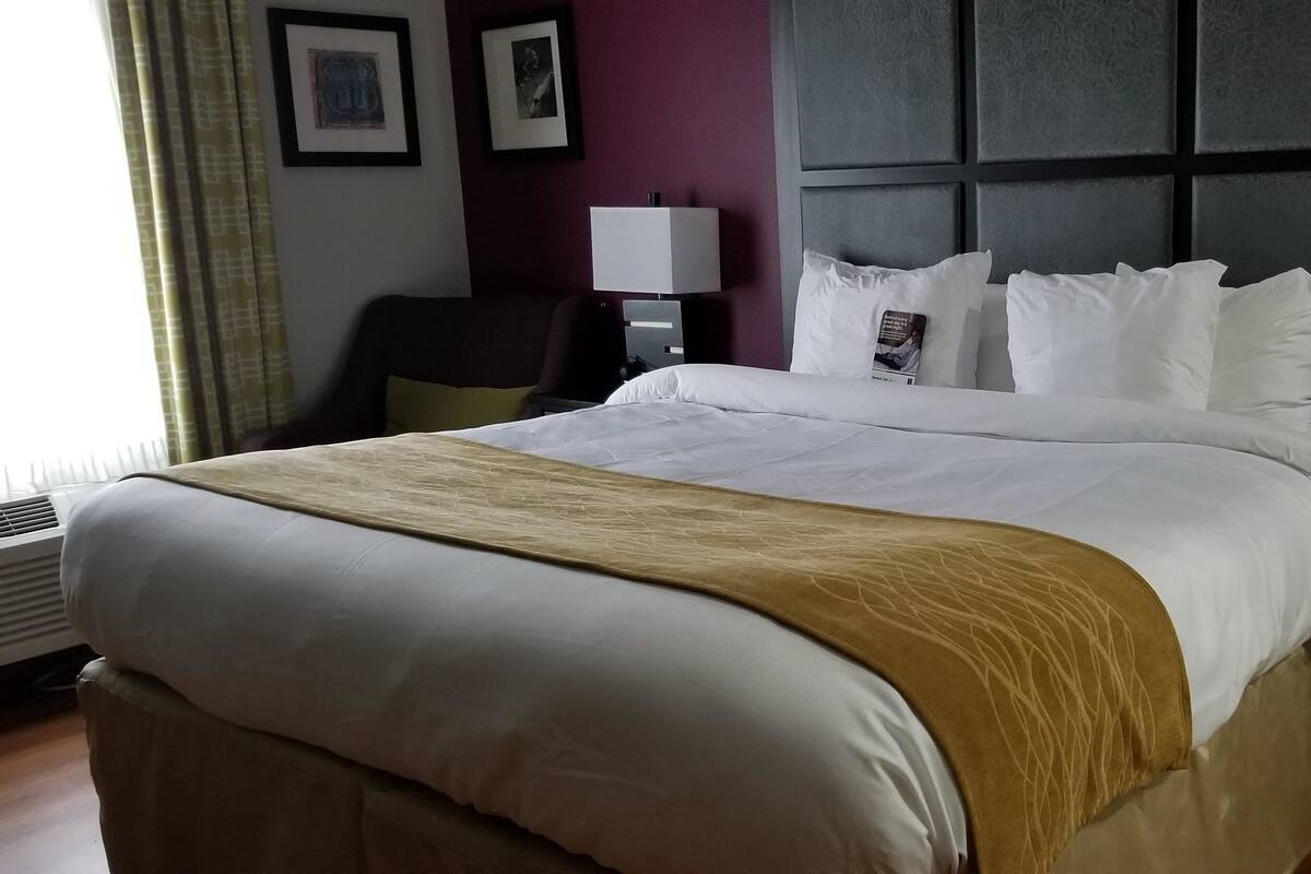 A hotel room with a large bed, a bedside table with a lamp, a chair, and a desk with a computer.