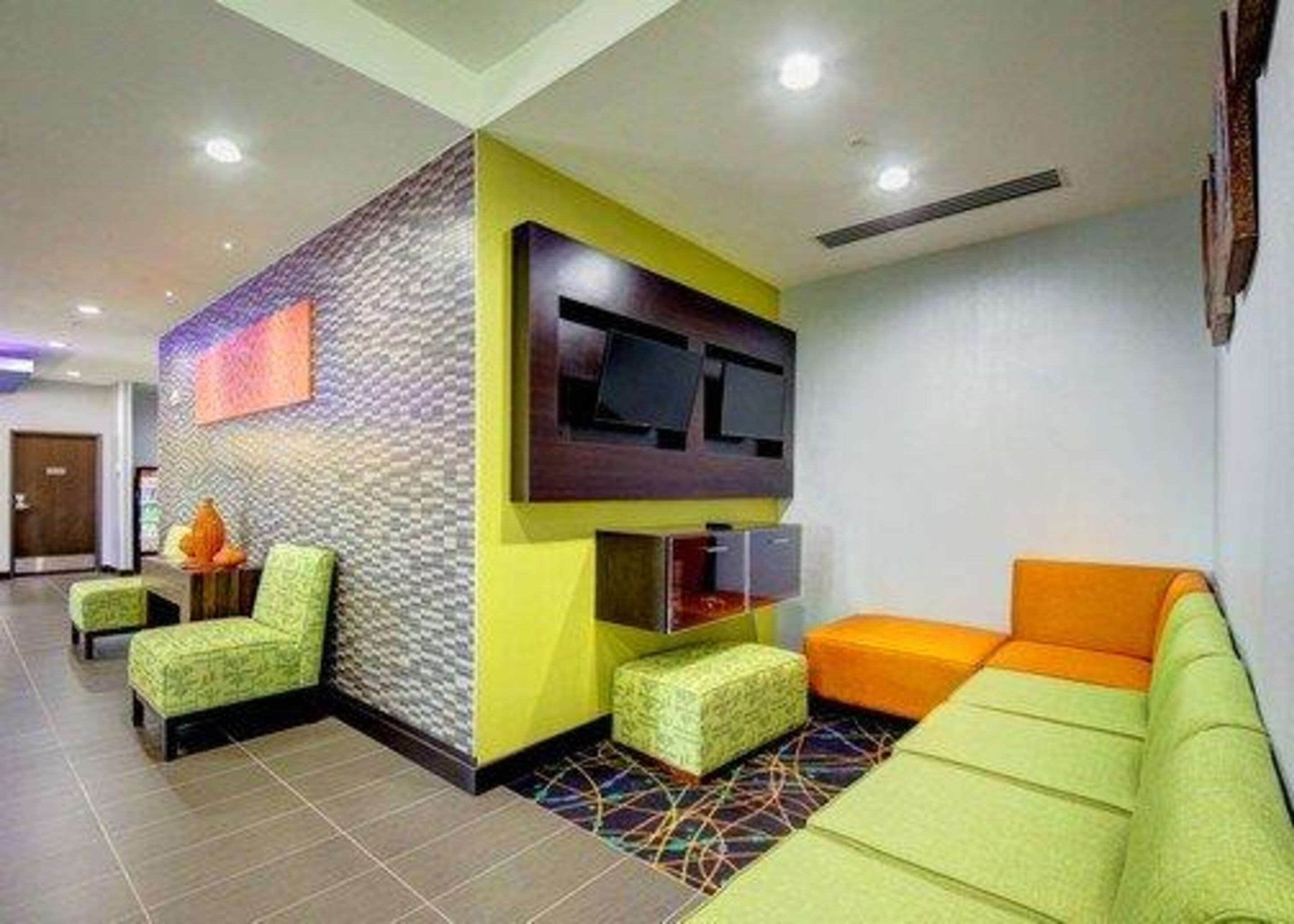 Un hall d’entrée moderne avec un mur décoré de motifs, un téléviseur à écran plat, des sièges verts et orange, et un sol carrelé.