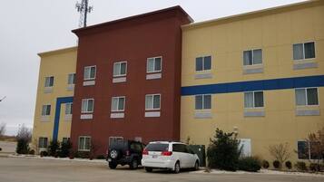 Un hôtel à plusieurs étages, doté d’une façade jaune et marron, de nombreuses fenêtres et d’un espace de stationnement où se trouvent deux voitures.