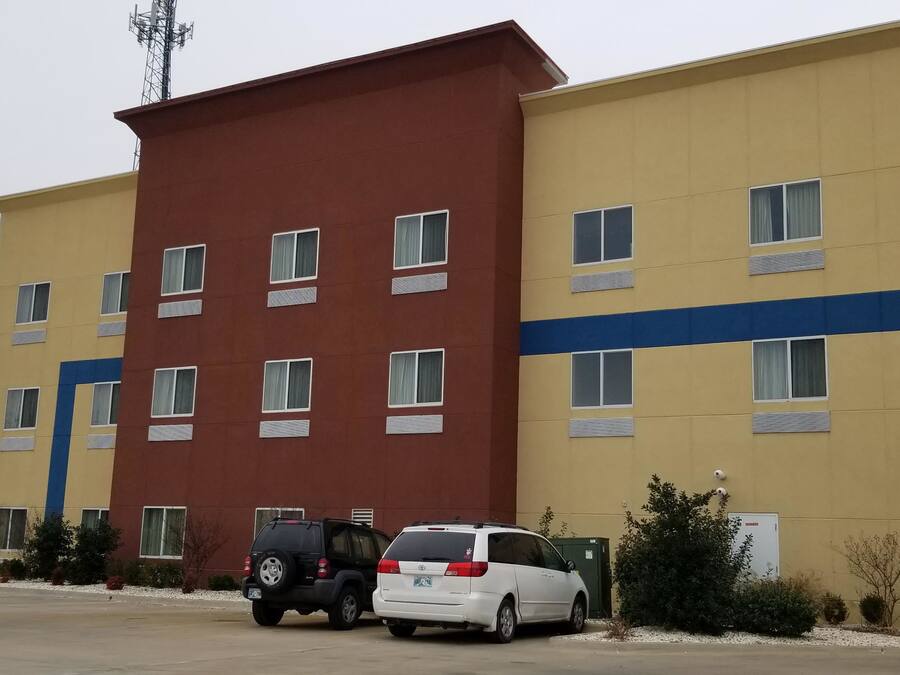 Un hôtel à plusieurs étages, doté d’une façade jaune et marron, de nombreuses fenêtres et d’un espace de stationnement où se trouvent deux voitures.
