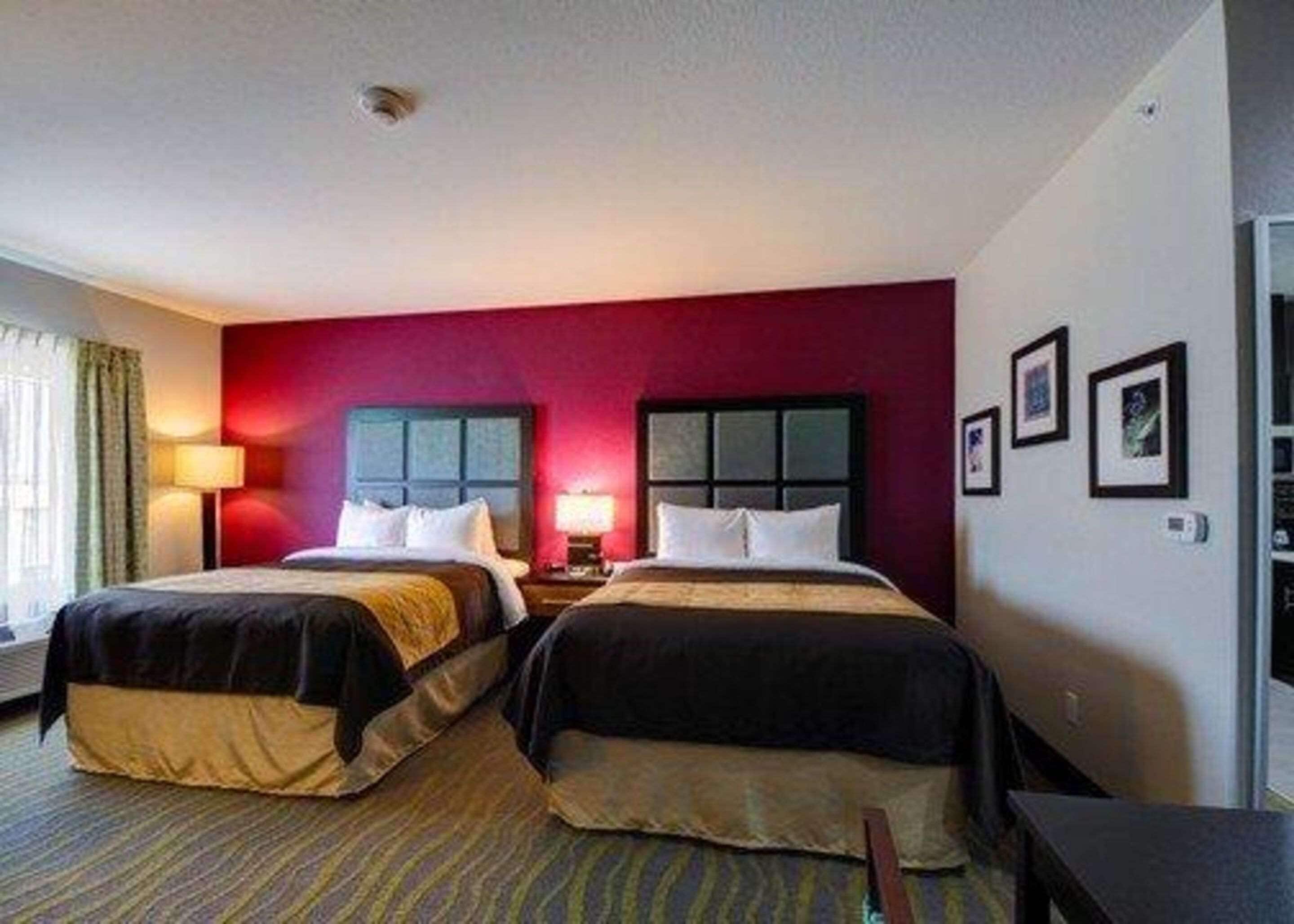 A hotel room with two beds, a red accent wall, a window with curtains, and a small table.