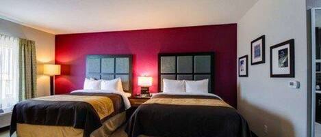 A hotel room with two beds, a red accent wall, a window with curtains, and a small table.