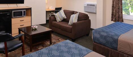 A hotel room with a sofa, two single beds, a desk with a flat-screen TV, a microwave, and a small table.
