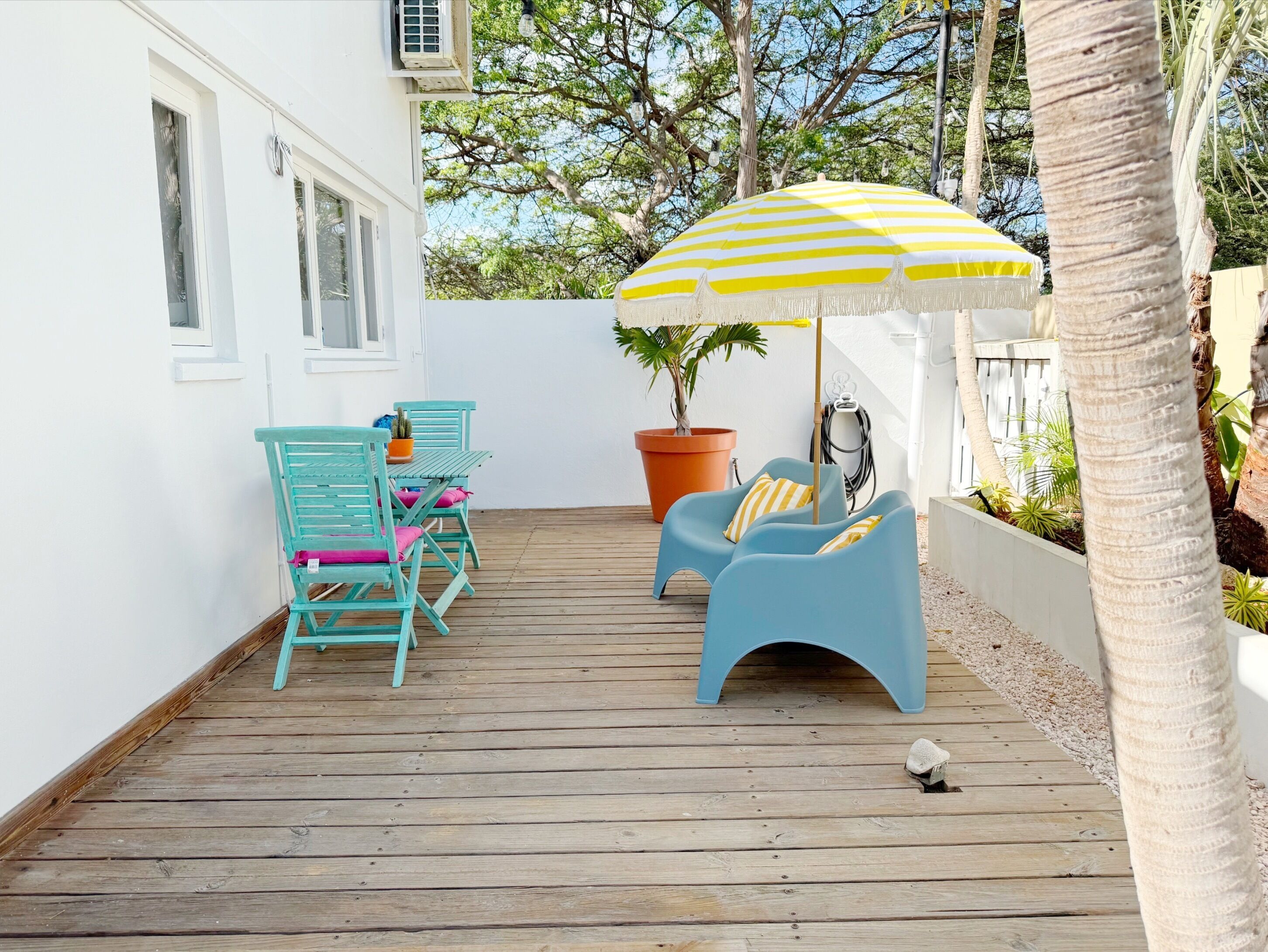 A wooden deck with turquoise chairs and a yellow-striped umbrella, surrounded by white walls and potted plants.