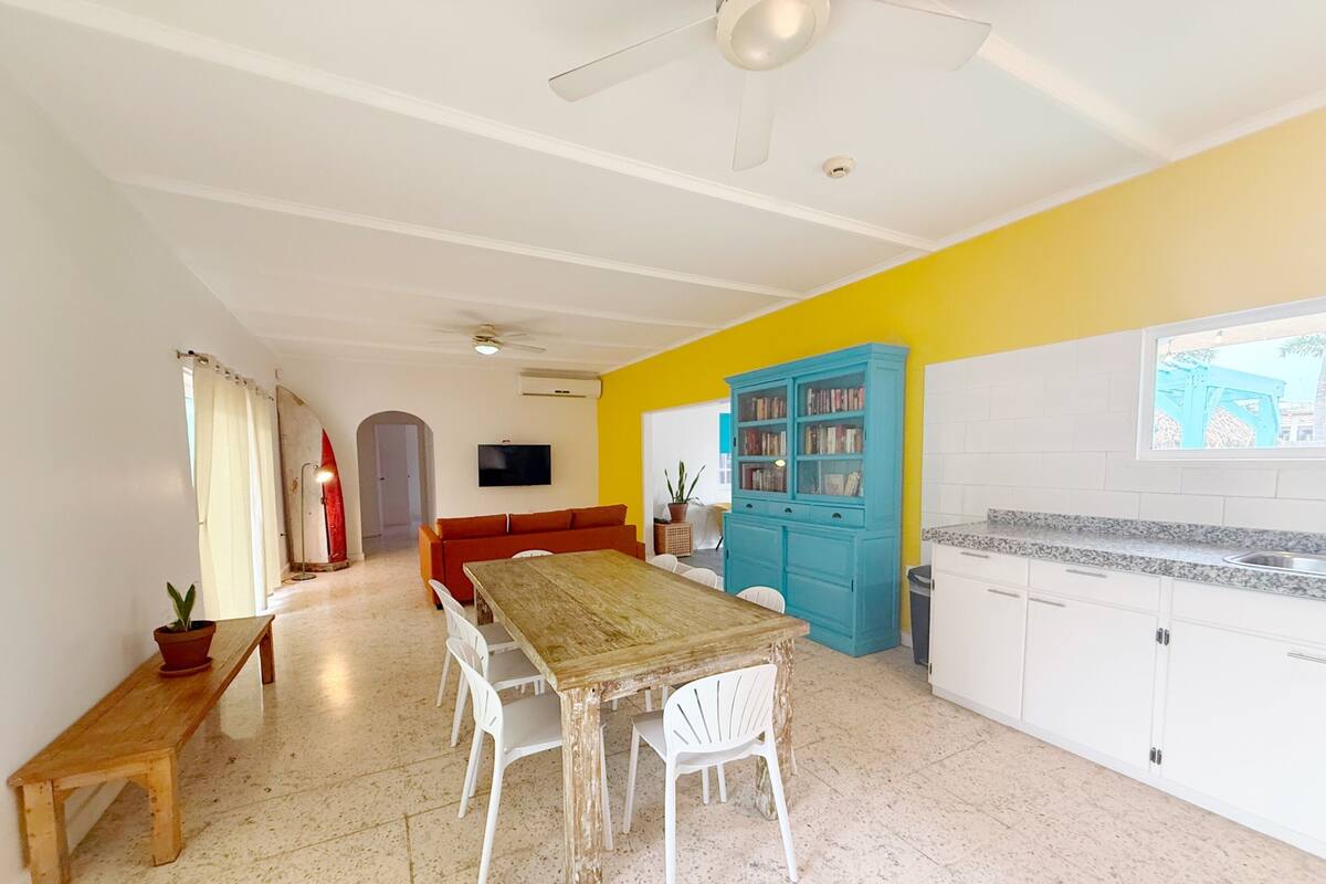 Amplia sala de estar con una mesa de comedor de madera, sillas blancas, un armario azul y una cocina con armarios blancos y encimera de granito.